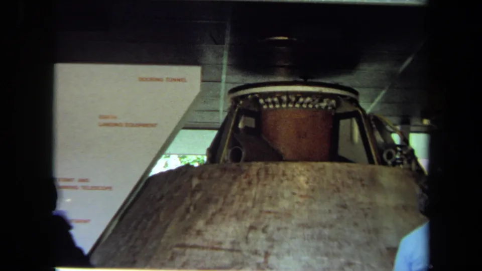 1970 California home-movie frame of a spacecraft display, partially framed by visitors and indoor exhibit signage.