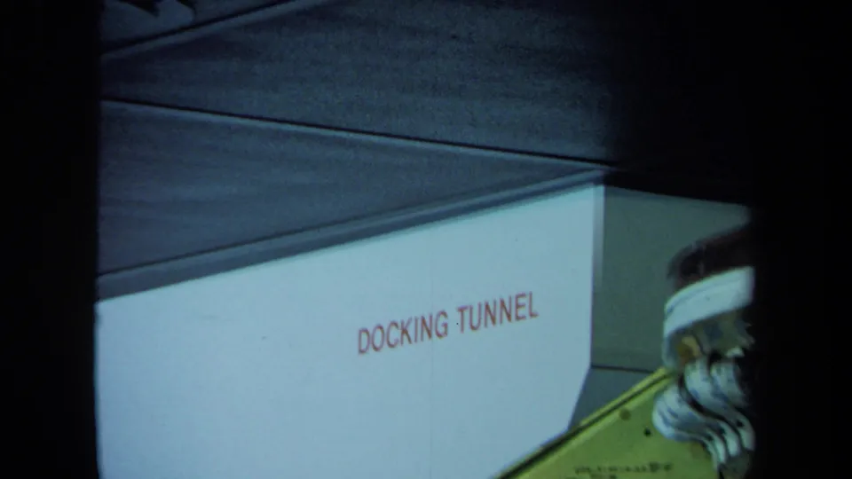 1970 California 8mm frame showing a wall marked DOCKING TUNNEL and part of a large machine under a dark ceiling.