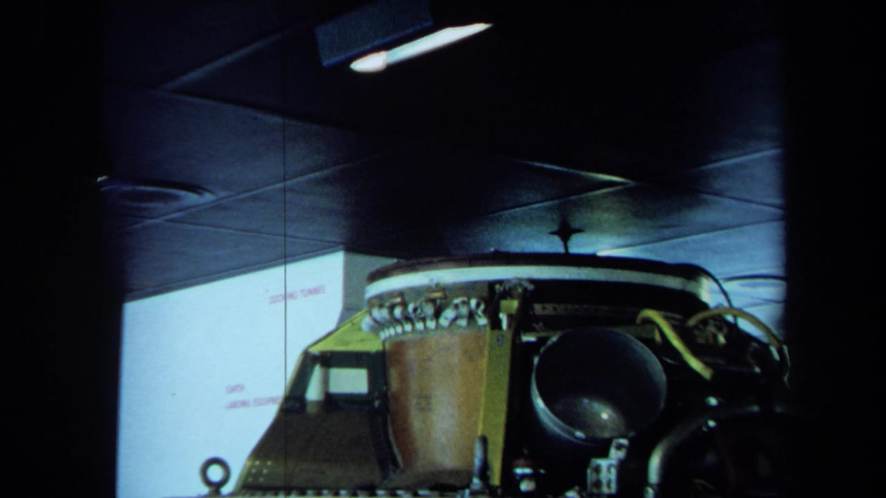 1970 California 8mm frame showing machinery, wiring and ceiling lights inside a dark docking tunnel space.