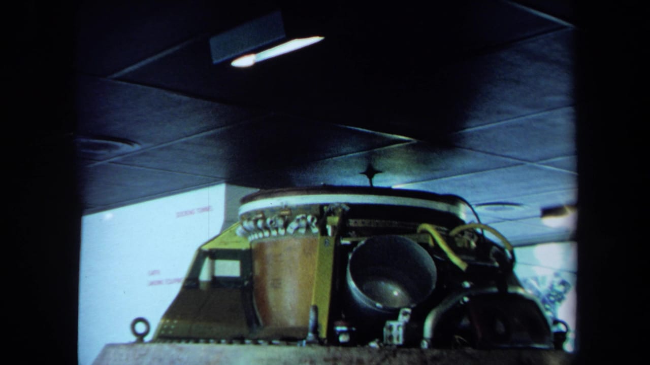 1970 California 8mm frame showing a spacecraft-like cockpit structure in a dim garage with controls, metal surfaces, and overhead lighting.