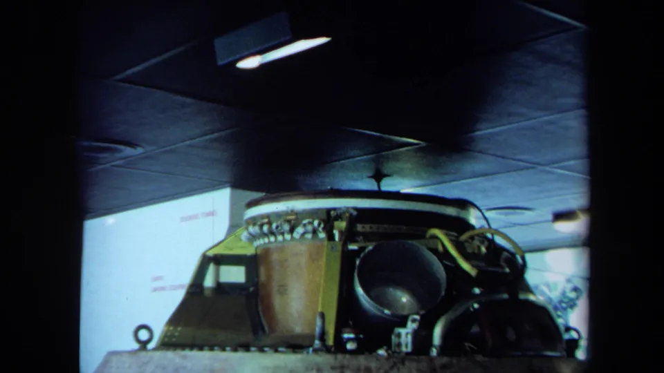 1970 California 8mm frame showing a spacecraft-like cockpit structure in a dim garage with controls, metal surfaces, and overhead lighting.