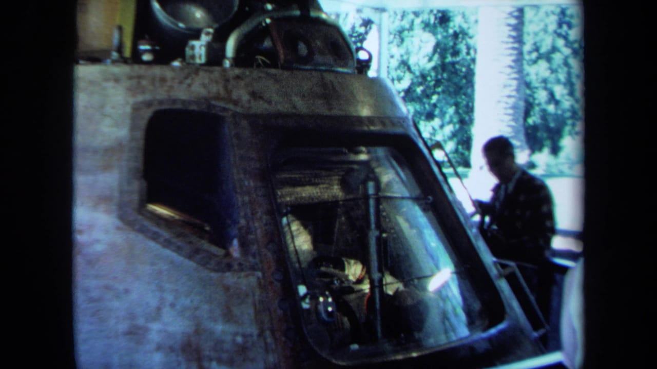 Man stands beside a large cockpit-like machine with a curved front window in a garage.