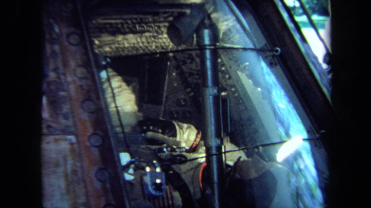 1970 California 8mm close-up of a spacecraft-like cockpit window with a suited, gloved occupant seated among controls and cables.