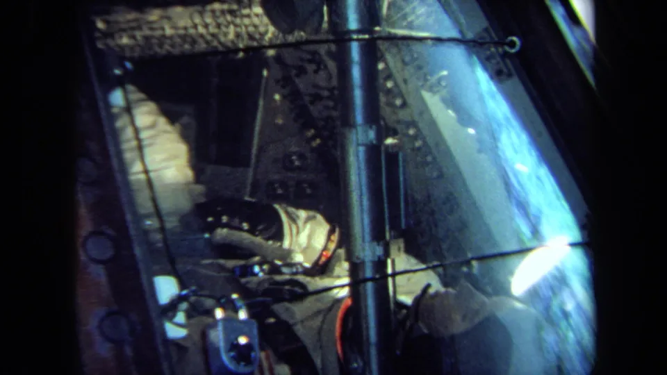 1970 California 8mm frame showing the cockpit-like interior with a suited occupant, gloved hand, window reflections, and metal hardware.