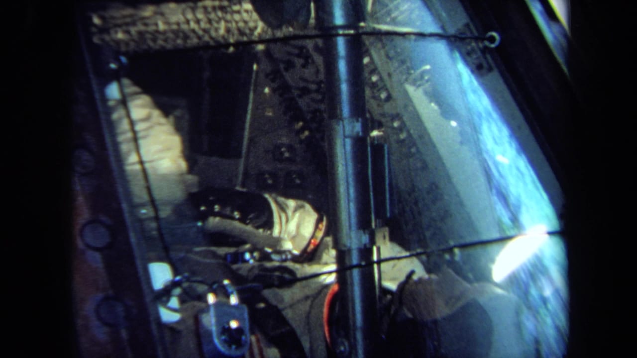 1970 California 8mm frame showing a white-suited figure inside a cramped spacecraft-style display seen through reflective glass.