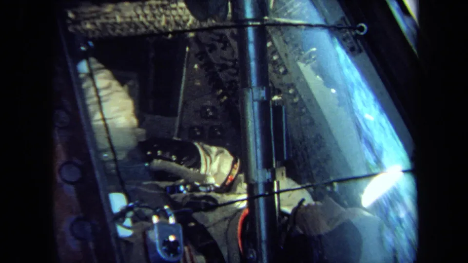 1970 California 8mm frame showing a white-suited figure inside a cramped spacecraft-style display seen through reflective glass.
