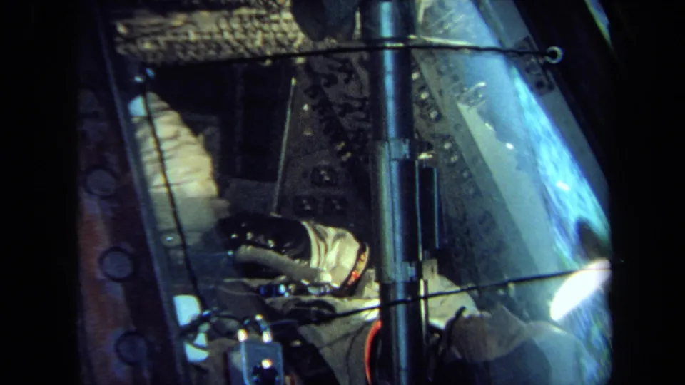 The white-suited figure remains visible inside the spacecraft-style training model, partly obscured by reflections and framing.
