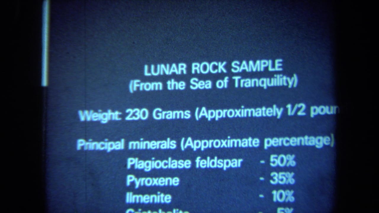 1970 California 8mm frame showing a projected informational slide with lunar rock sample weight and mineral percentages in blue light.