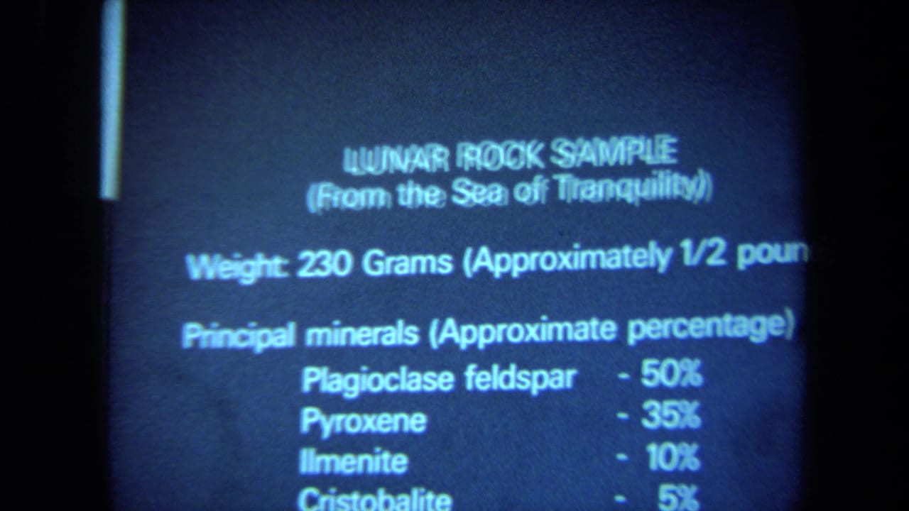 California 1970 8mm frame showing a museum label for a lunar rock sample, with mineral percentages partly readable in blue light.