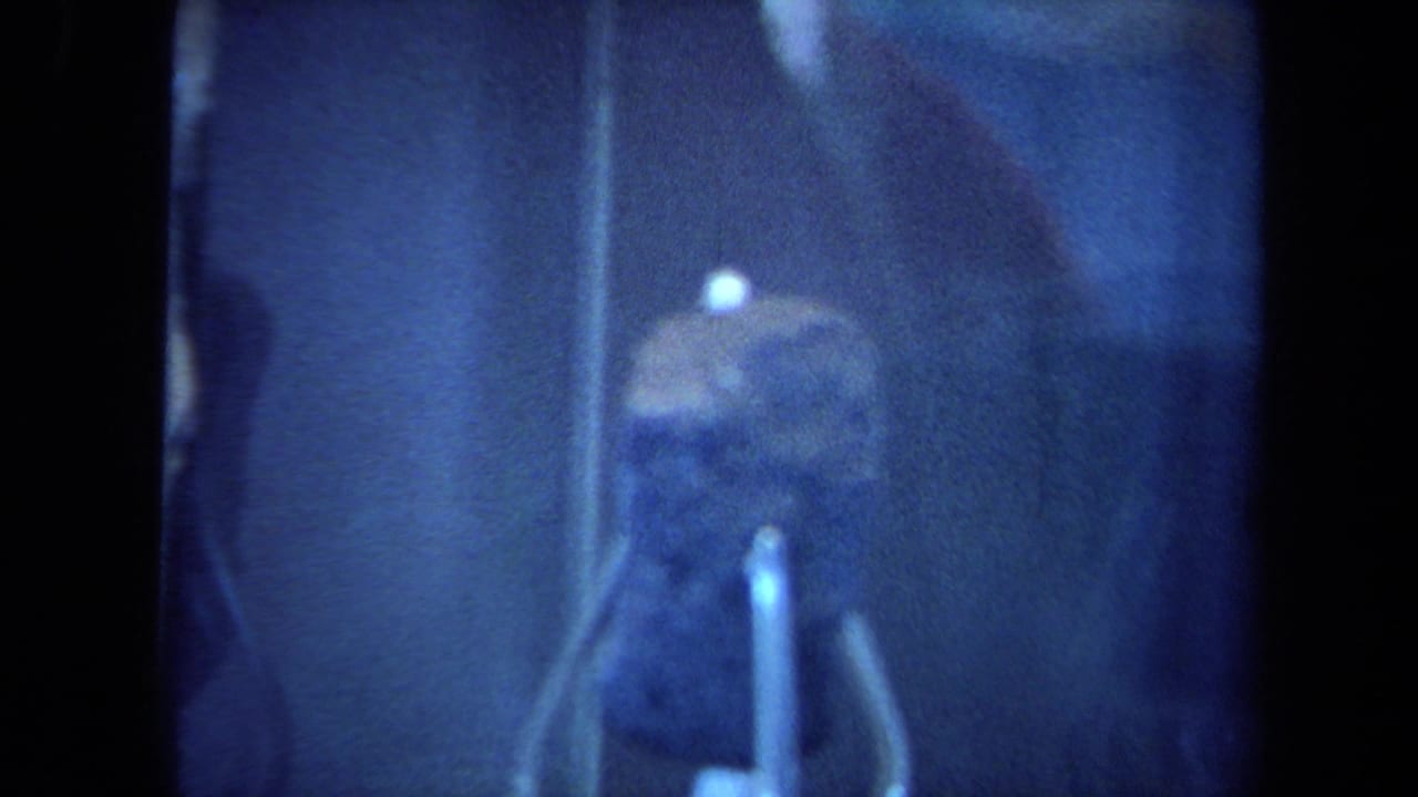 Dark rock specimen inside a glass display case with support pins visible.
