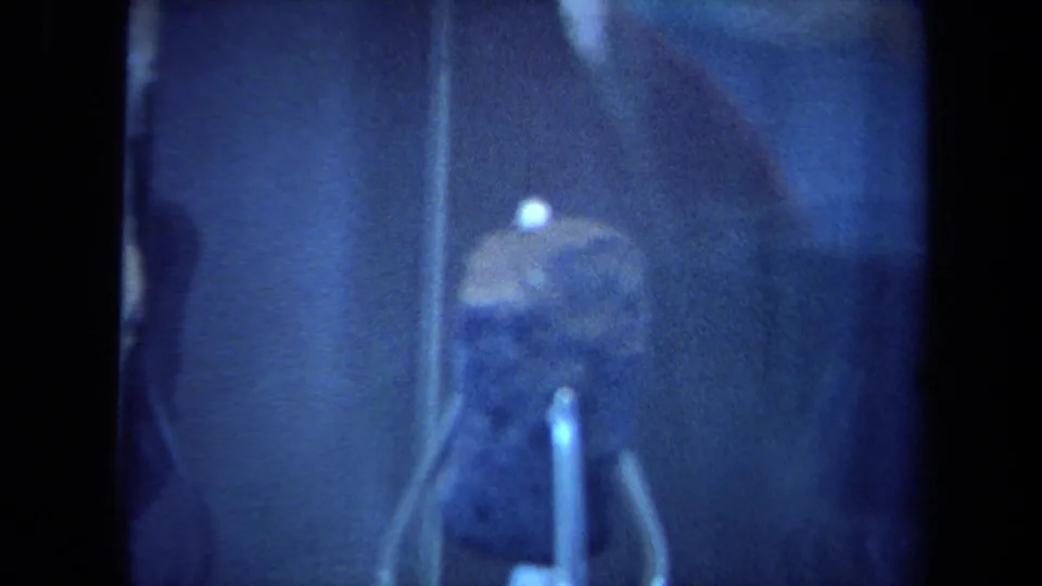 California 1970 8mm frame showing a lunar rock specimen behind glass, softly focused under blue museum lighting.