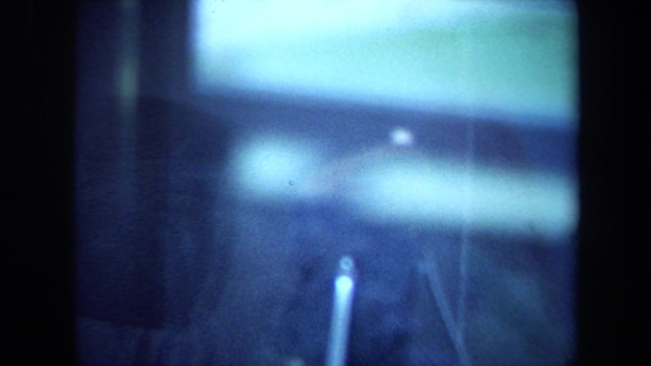 California 1970 8mm frame with a blurred museum-case view of the lunar sample area, including glare, support pins and blue light.