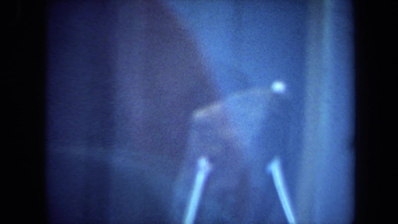 California 1970 8mm frame showing the lunar rock sample behind glass, with support pins, reflections and a blue cast from exhibit lighting.