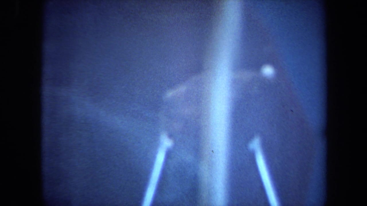 Soft, blue-tinted 1970 California 8mm frame showing a rock-like illuminated display with bright reflections and heavy grain.