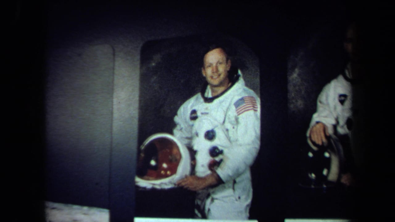 1970 California 8mm frame showing a smiling man in a white NASA-style spacesuit holding a helmet, with another suited figure partly visible.