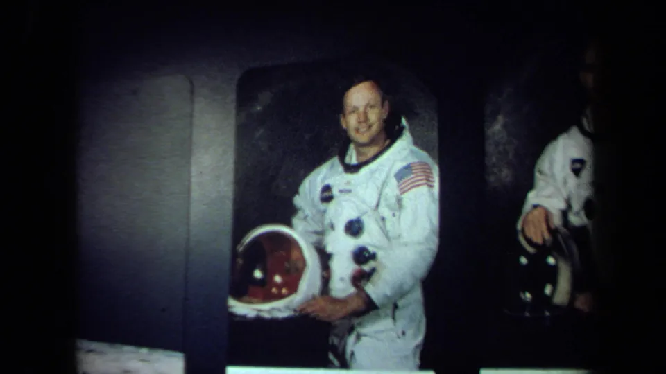 1970 California 8mm frame showing a smiling man in a white NASA-style spacesuit holding a helmet, with another suited figure partly visible.