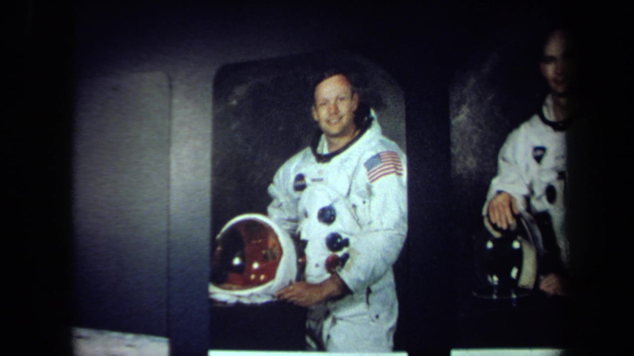In 1970 California footage, an astronaut in a white spacesuit with a flag patch smiles and holds a helmet against a dark studio-like background.