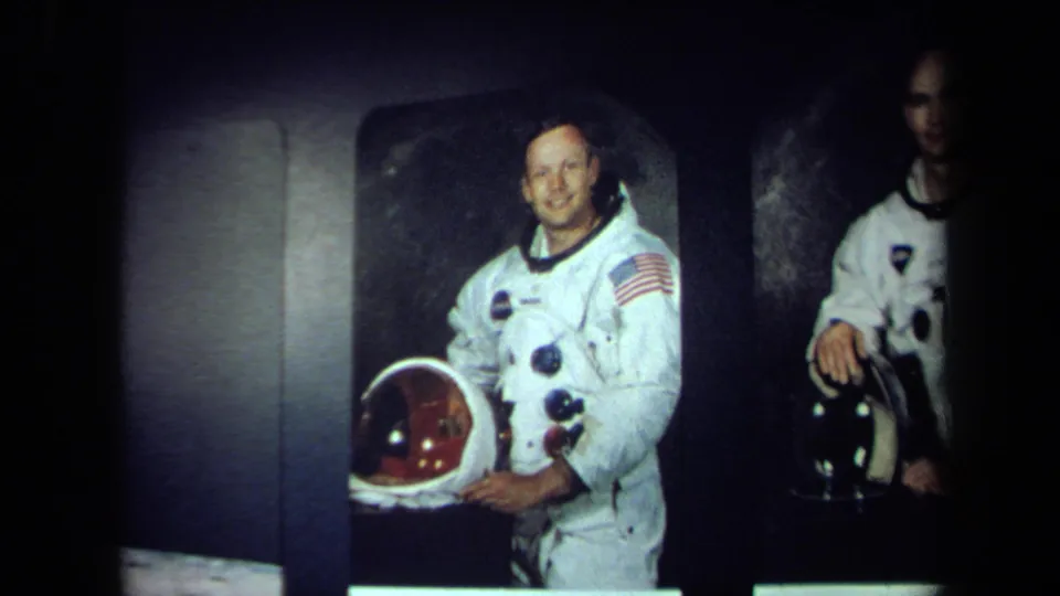 In 1970 California footage, an astronaut in a white spacesuit with a flag patch smiles and holds a helmet against a dark studio-like background.