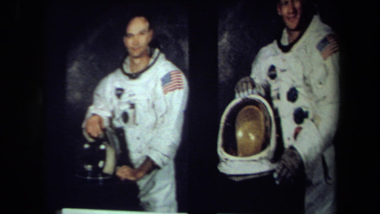 An astronaut in a white suit holds a dark helmet in the foreground, with another suited man and a reflective visor helmet nearby.