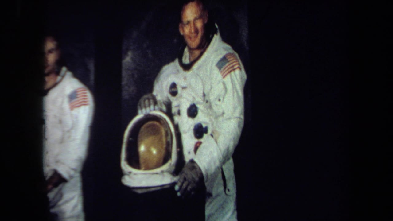 A smiling astronaut in a white spacesuit with a flag patch stands beside a helmet with a gold visor in 1970 California archival footage.
