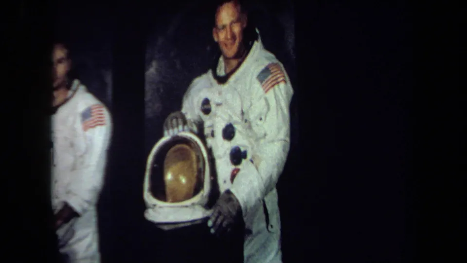 A smiling astronaut in a white spacesuit with a flag patch stands beside a helmet with a gold visor in 1970 California archival footage.