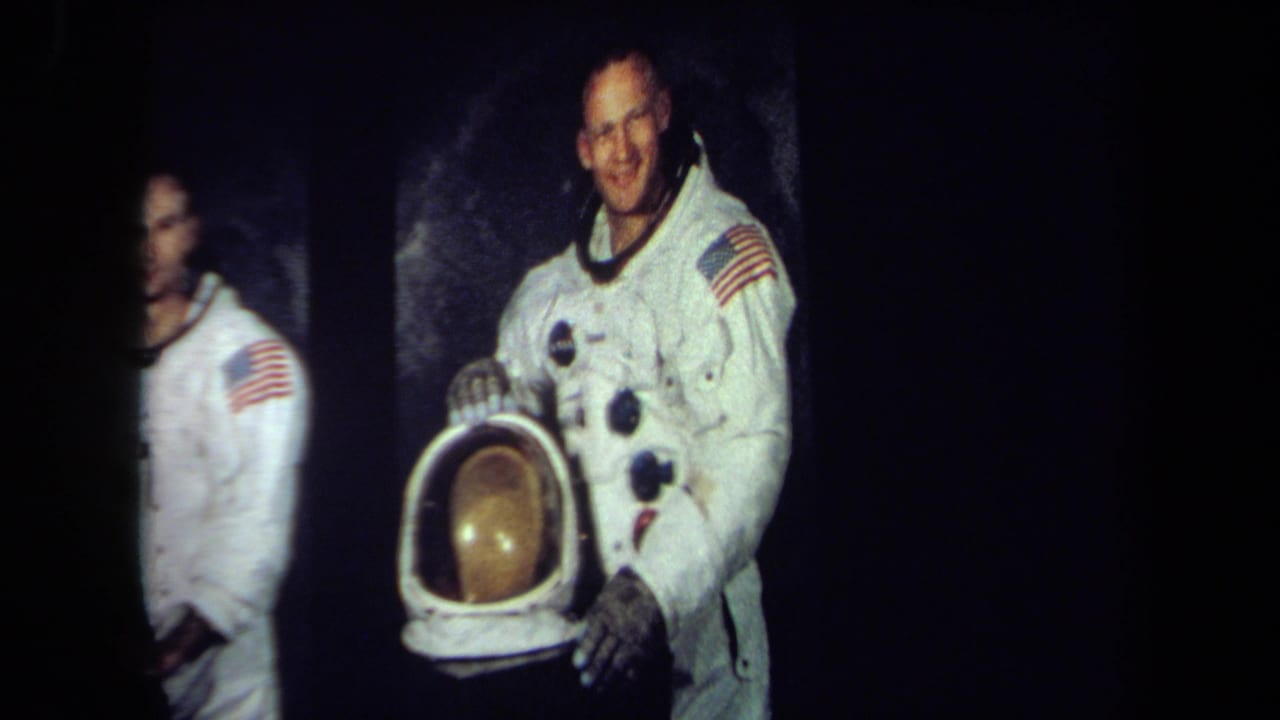 Two astronauts in white spacesuits stand against a dark background, one holding a helmet.