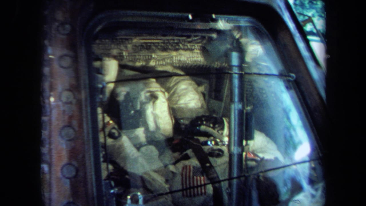 California 1970 8mm view of a spacecraft cockpit display with white-suited astronauts partly obscured by reflective glass.