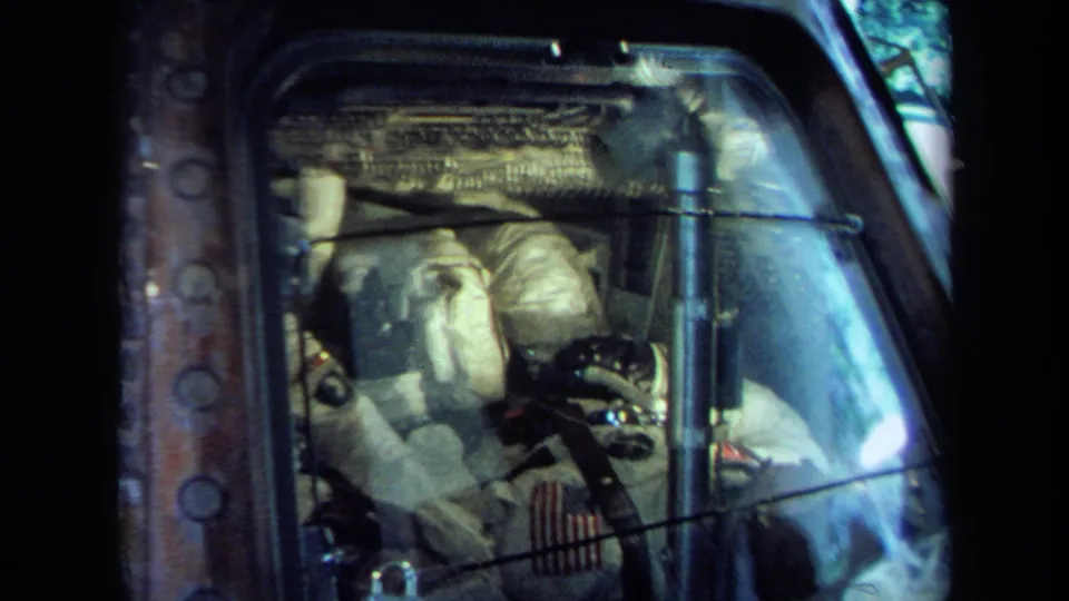 California 1970 8mm view of a spacecraft cockpit display with white-suited astronauts partly obscured by reflective glass.