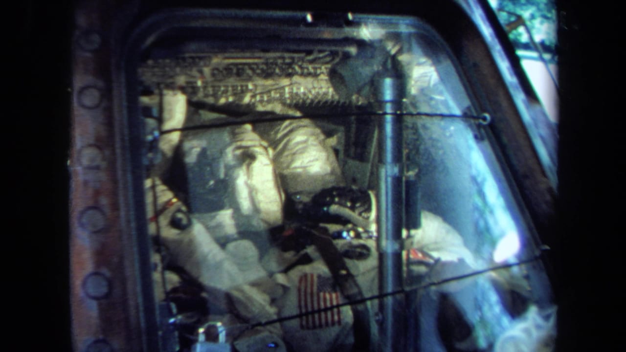 A spacecraft cockpit display in 1970 California footage shows suited figures behind glass with panels, wiring, and reflections.