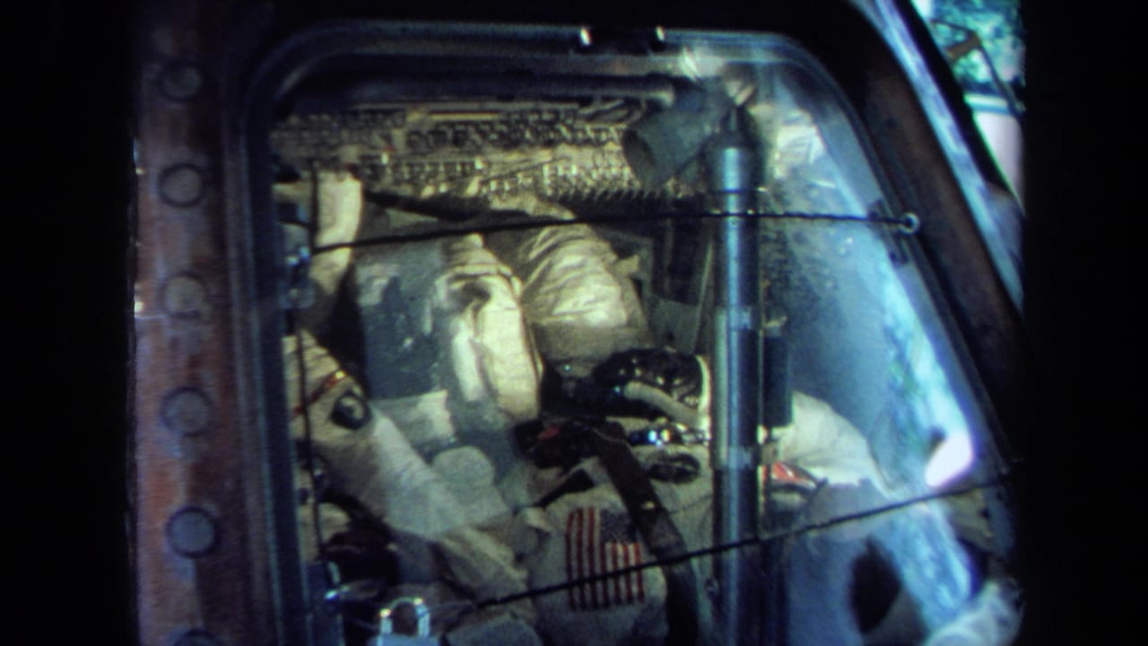 1970 California 8mm frame showing a white-suited astronaut figure inside a capsule, viewed through reflective glass with panels and wiring visible.