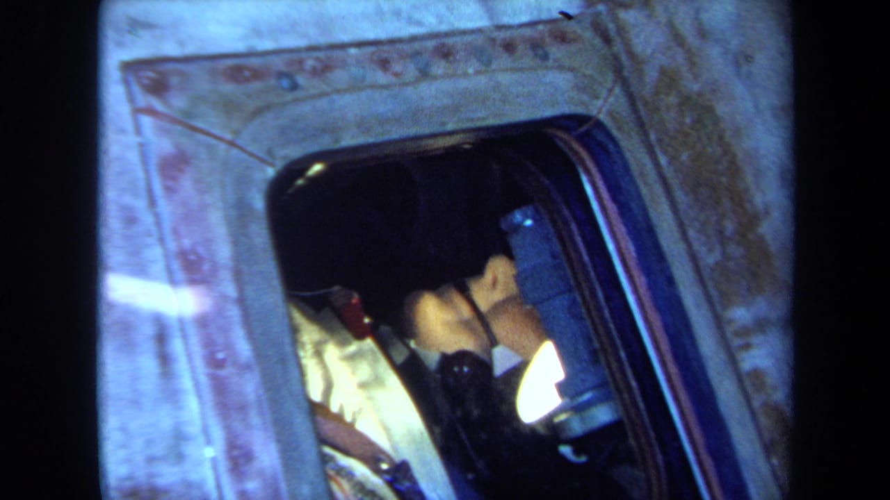 1970 California home-movie frame of a white-suited astronaut figure viewed through a metal capsule window, with raised arms and reflective glass.