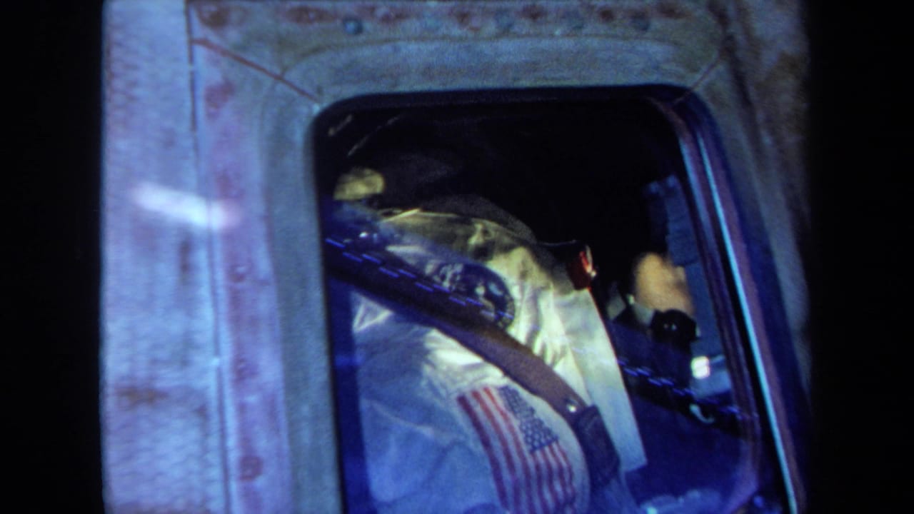 A white astronaut suit with an American flag patch is visible through the capsule window, with straps and blue-tinted reflections across the frame.