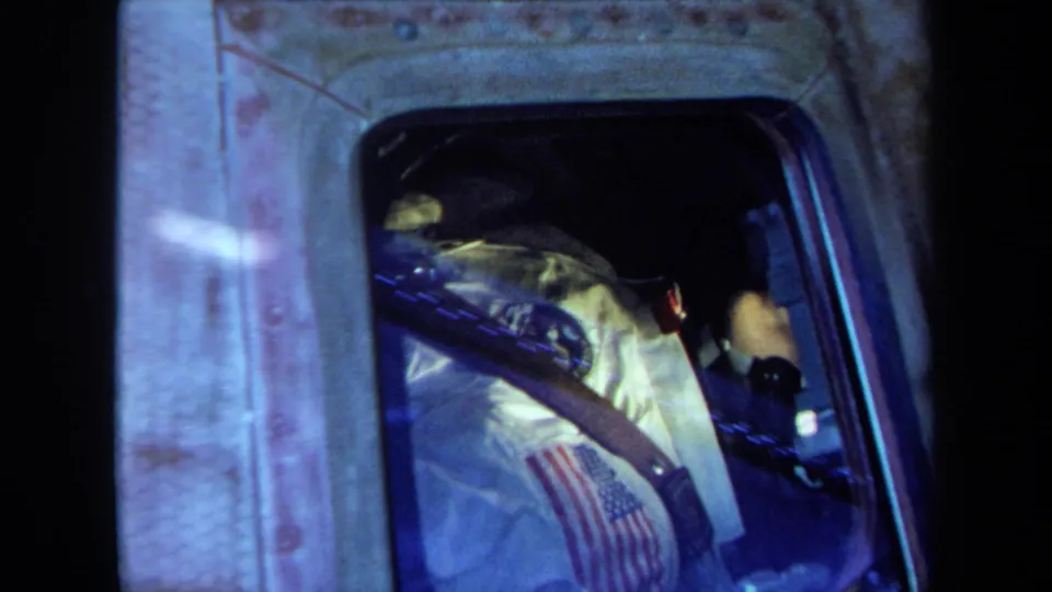 A white astronaut suit with an American flag patch is visible through the capsule window, with straps and blue-tinted reflections across the frame.
