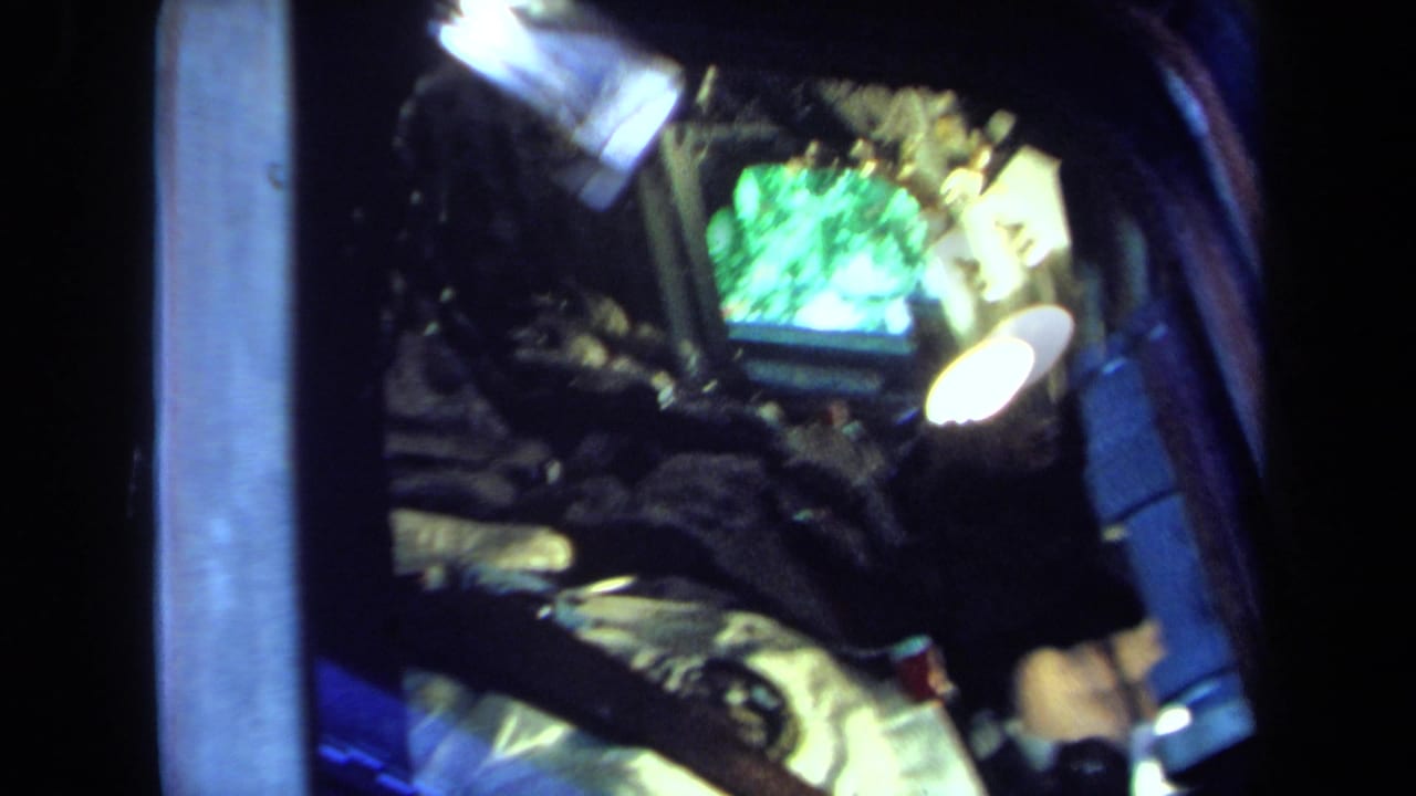 Dim 8mm view into a spacecraft-style cabin with a seated astronaut figure, bright lamps, straps, and a green-tinted display.