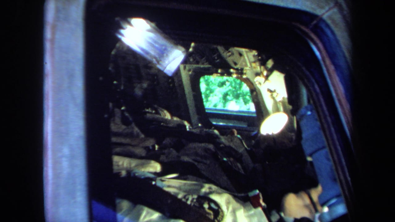 Handheld 8mm frame looking into a spacecraft display with illuminated equipment, a green screen, and the suited figure partly visible.