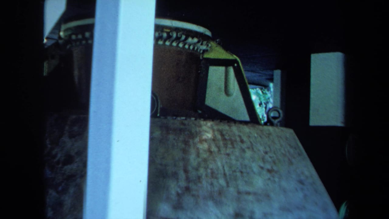 1970s California home-movie frame showing a large concrete-like slab near a dark bus or vehicle, with white posts and rounded window shapes.