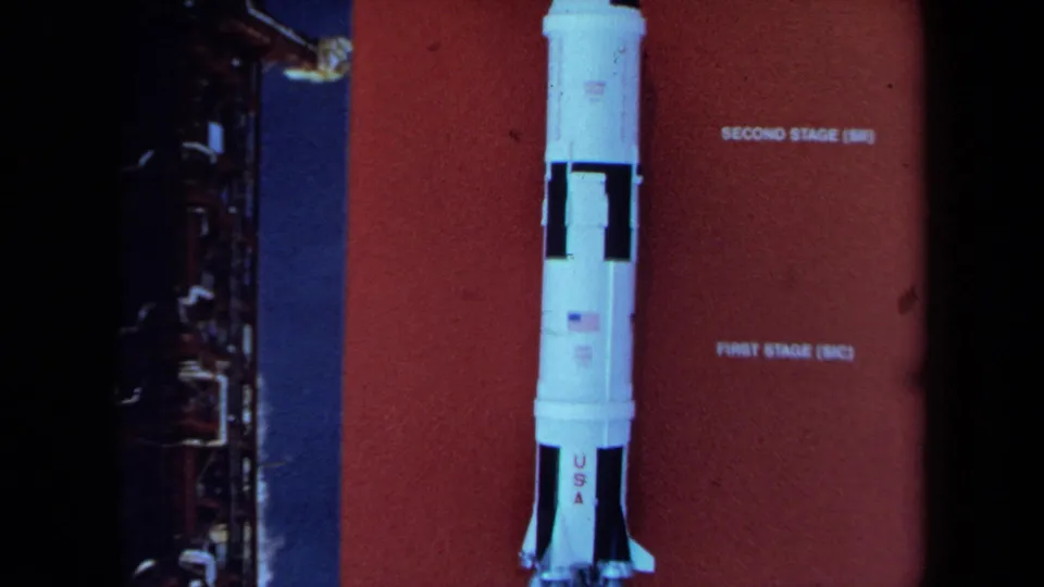 1970 California 8mm frame showing a white NASA-style rocket model against a red stage-labeled display panel.