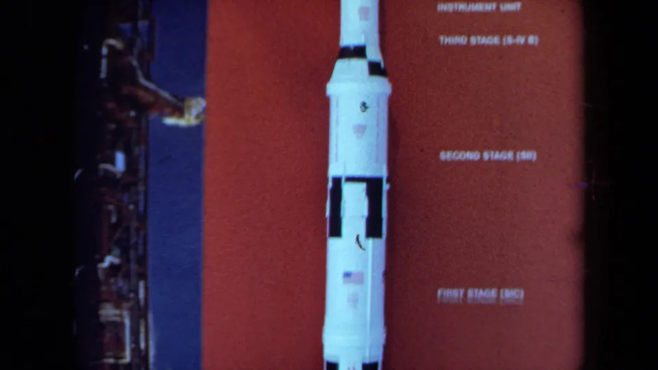 A white rocket model appears beside labels for its stages on a red educational display, filmed in soft 8mm color.
