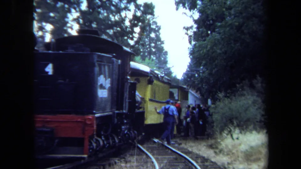 1970 California 8mm frame showing a yellow passenger train beside wooded tracks as passengers stand near the cars.