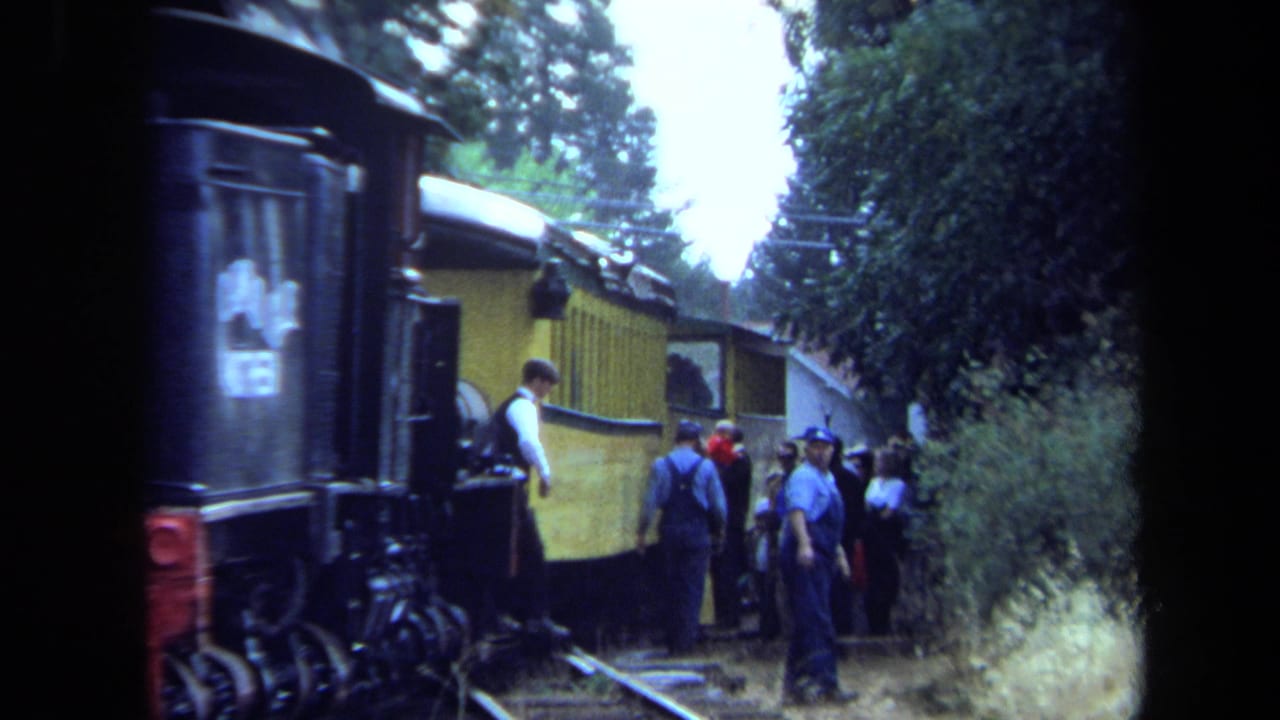 Crew member and passengers beside a yellow train car in a wooded area.