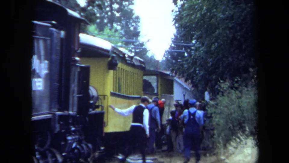 Passengers in casual 1970s clothing move around a yellow railcar during a wooded train stop in California.