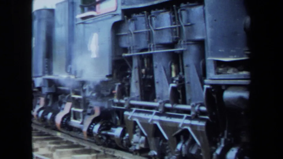 1970 8mm California home-movie frame showing a close side view of a steam locomotive's wheels, rods, and lower body as steam drifts near the track.