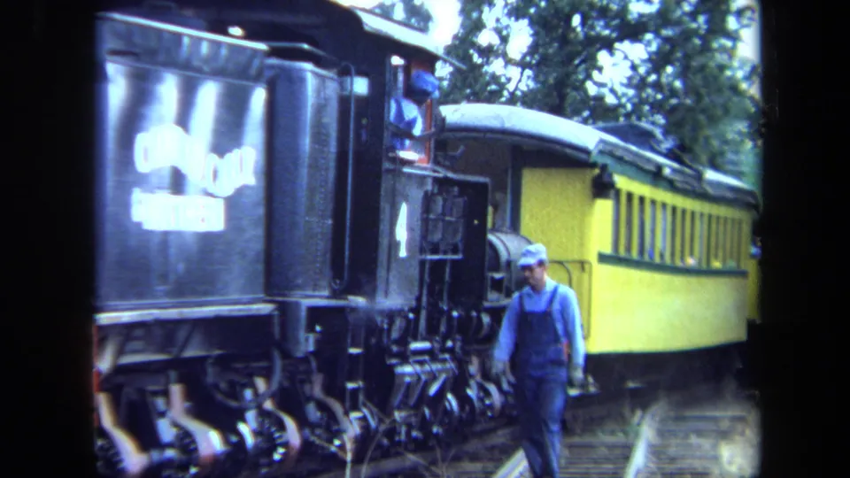1970 California home-movie frame showing an engineer walking beside a black steam locomotive marked with number 4 and a yellow passenger car.
