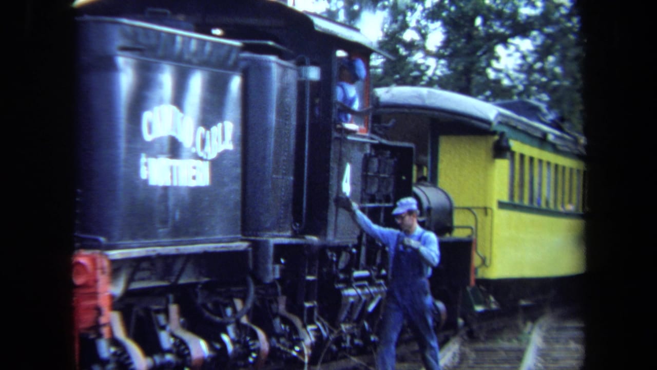 Worker in blue overalls steps onto a black steam locomotive beside a yellow passenger car.