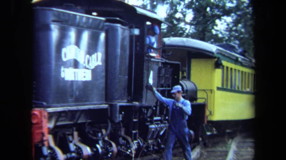 1970 California 8mm frame showing a rail worker boarding a black steam locomotive with white side lettering near a yellow passenger car.