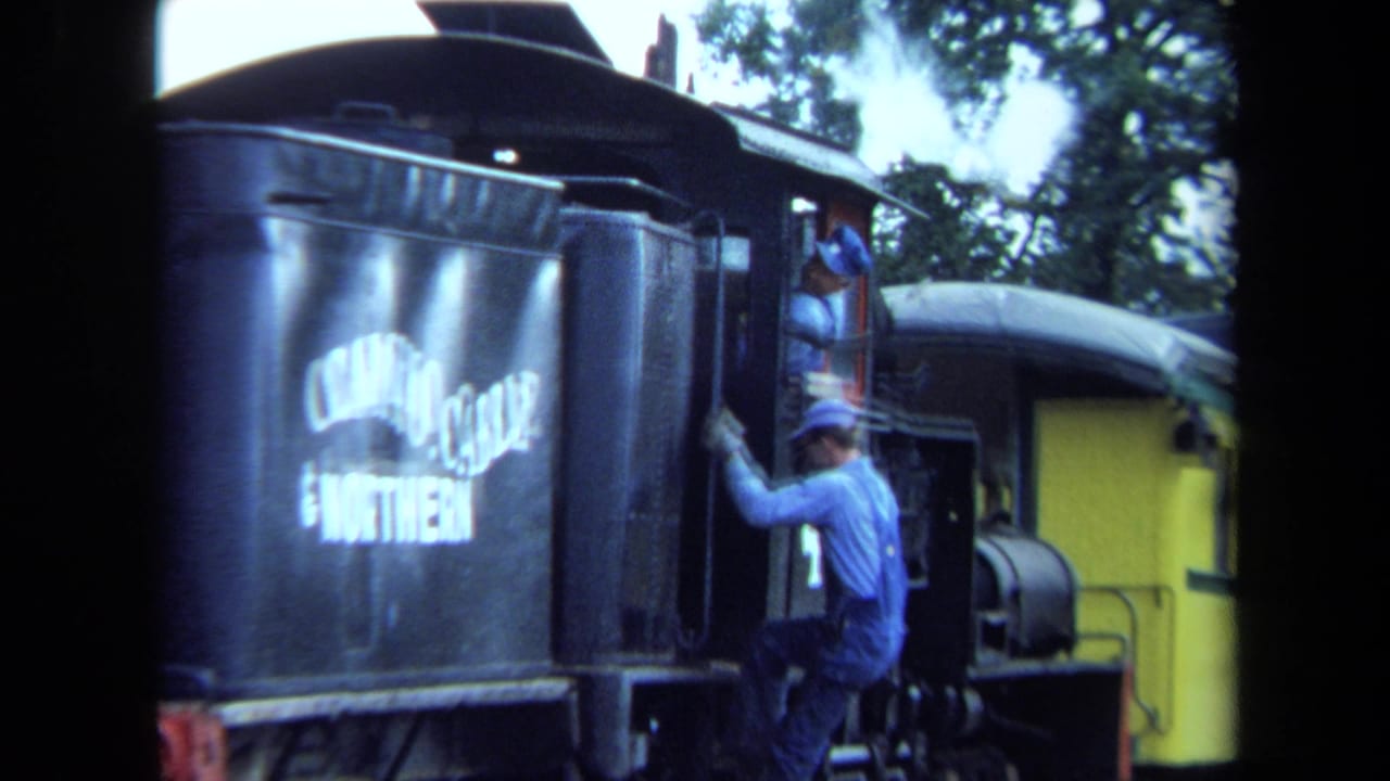 Close 8mm view of a worker climbing aboard a black steam locomotive in California, with a yellow passenger car visible behind.
