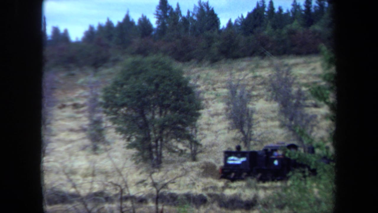 Small black steam train with visible riders or figures moves along a rural California hillside in 1970 home-movie footage.
