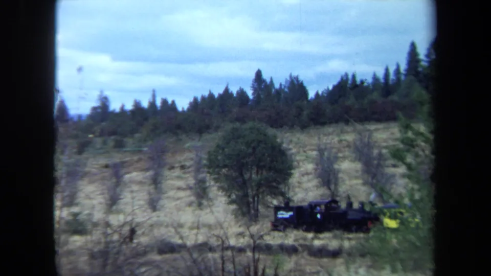 1970 California home-movie frame of a black steam train pulling a yellow passenger car beneath a wooded ridge.
