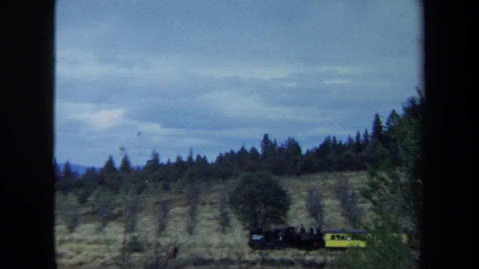Distant 1970 California 8mm view of a small steam train and yellow car traveling across a rural hillside.