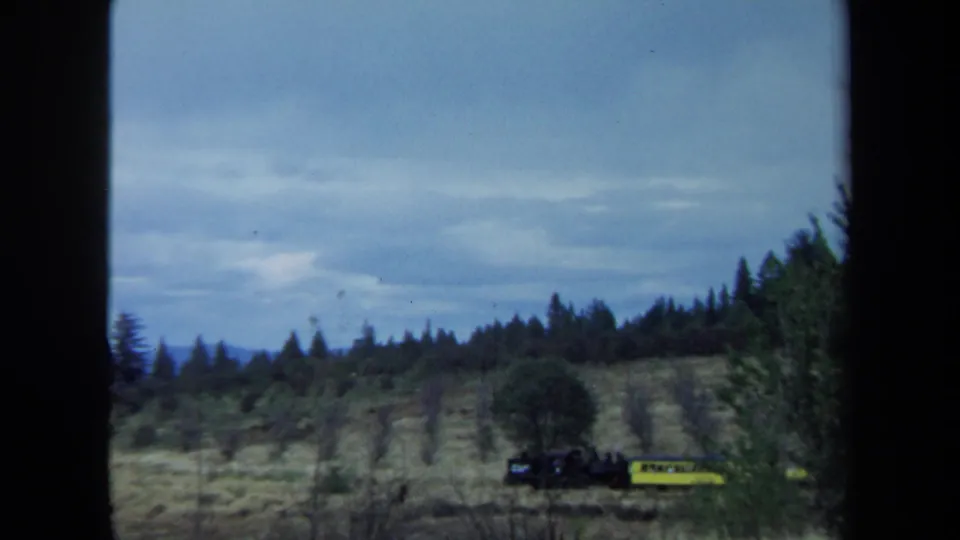 In 1970 California 8mm footage, a small train with a yellow car passes through dry grass below wooded hills and an overcast sky.